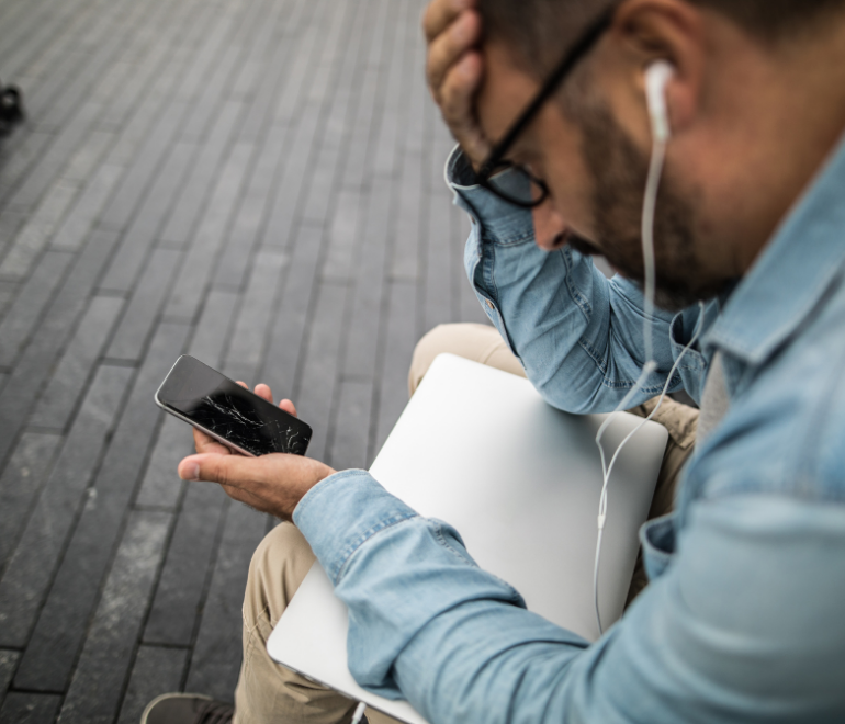 Sad man with apple ear buds and macbook computer holding a broken iphone with cracked screen