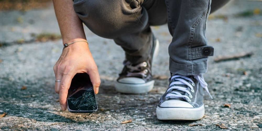 below-the-knee photo of someone bending down to pick up a smartphone with a cracked screen.