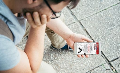 A man sitting on a bench looking forlornly at his smart phone with cracked glass